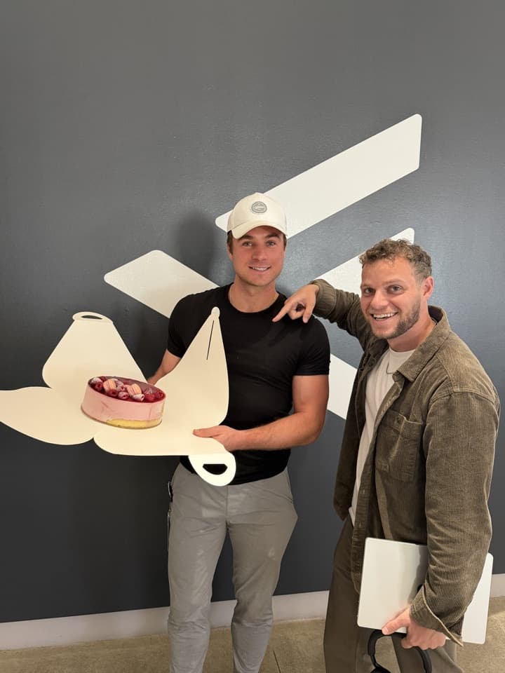 Alex Carrabre at F.inc holding a pink cheesecake coldcake on an F-shaped board, F.inc graphic wall behind him