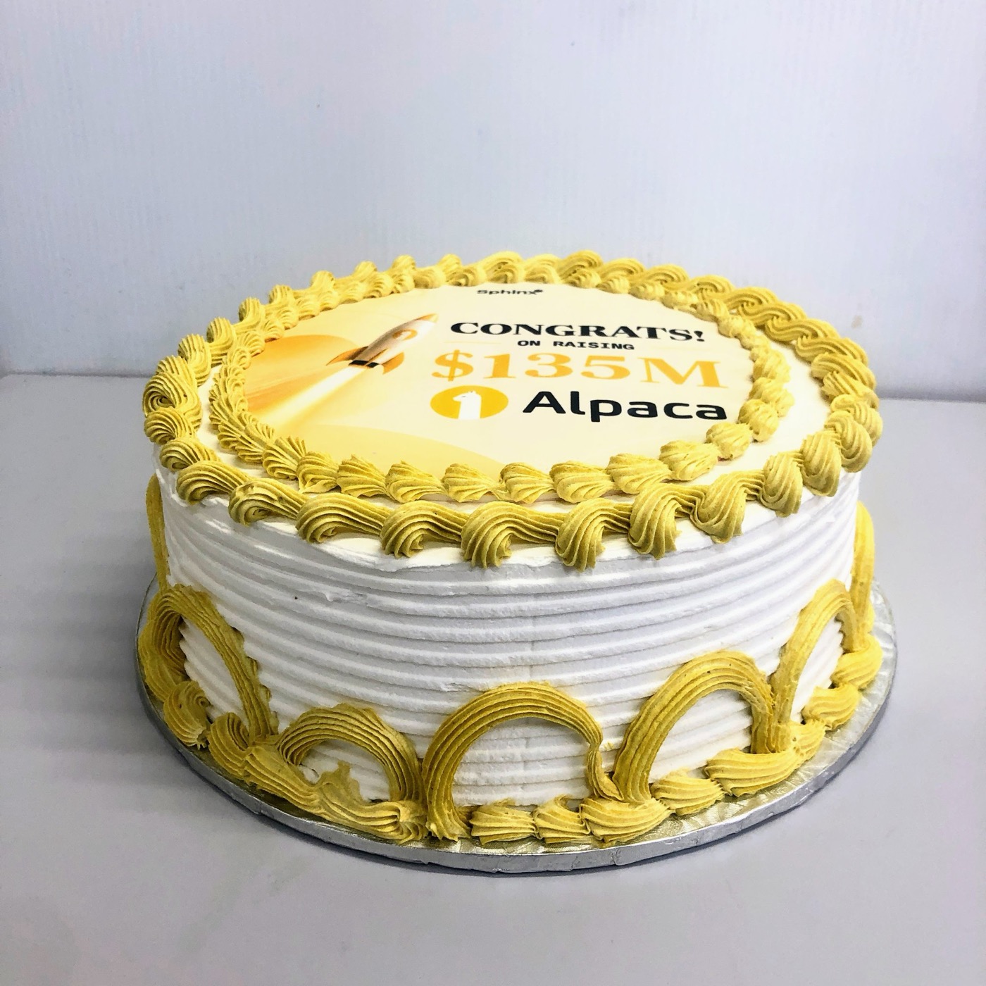 A cake with an edible printed top reading "Congrats on raising $135M" above a company logo, finished with piped yellow buttercream.